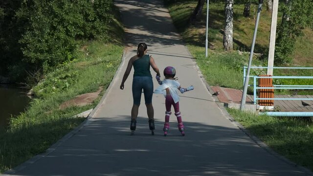 Little Girl in Protection Pads and Colorful Helmet Falling while Roller Skating with Mother in a Park. Slow Motion. Childhood, Summer Activities and Healthy Lifestyle Concept