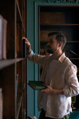 A man in a light linen shirt stands in front of a bookshelf, chooses something and holds a green book in his hand