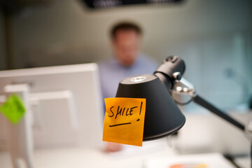 Closeup of sticky note with smile text stuck on desk lamp at illuminated creative office