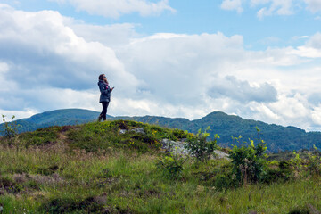Girl with cell phone in her hand are standing on a hill looking at the mountain view. Tourist, technology and travelling concept.
