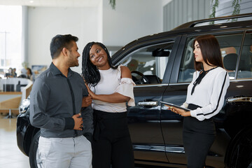 A beautiful couple smiles while looking at a new car in a car dealership with a seller. An agent demonstrates the new features of a luxury car to an African-American happy couple at a car dealership.