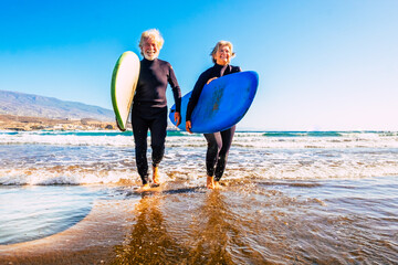 two old and mature people having fun and enjoying their vacations outdoors at the beach wearing wetsuits and holding a surfboard to go surfing in the water with waves - active senior smiling and enjoy - obrazy, fototapety, plakaty