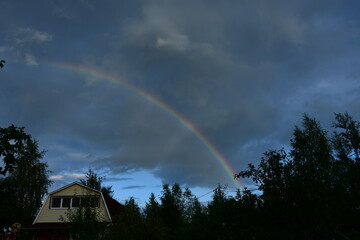 Summer evening at the dacha. A rainbow over the house in a frowning sky.