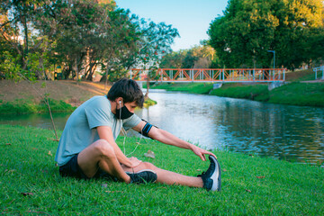 Fototapeta premium A young man doing stretching in a green park before starting to exercise in sportswear and mask.