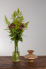 Glass vase with bouquet of beautiful flowers near wood vase over wood table on white wall background