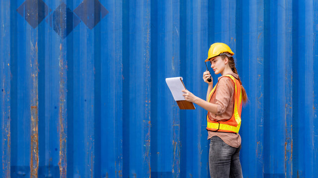 Confident inspector standing in front of cargo container in shipping yard. Smart female engineer, dock worker wear hard hat using clipboard and radio to command transportation. Concept of logistics