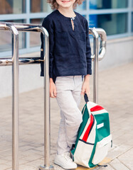 Schoolboy boy in light pants and a dark shirt posing with a backpack in his hands
