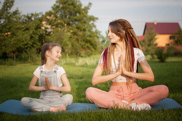 Young mother with dreadlocks and little daughter are doing yoga exercises in lotus position on grass in the park at the day time. Concept of friendly family and of summer vacation.
