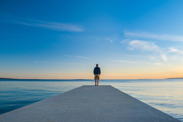 Man in hoodie and short cargo pants standing on dock and looking on sea horizon, island od Pag,...