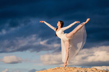 young ballerina in a light long white dress stands in an arabesque like a bird, her skirt develops...