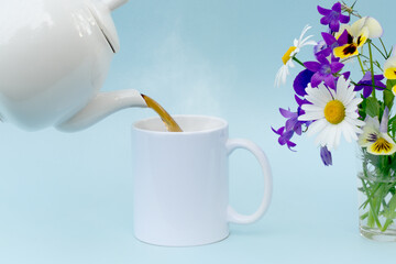 Hot tea from a white teapot is poured into a white mug, no logos. next to a bouquet of wildflowers. Blue background. Soft focus, close-up, horizontal photo. Concept - layout for advertising, postcards