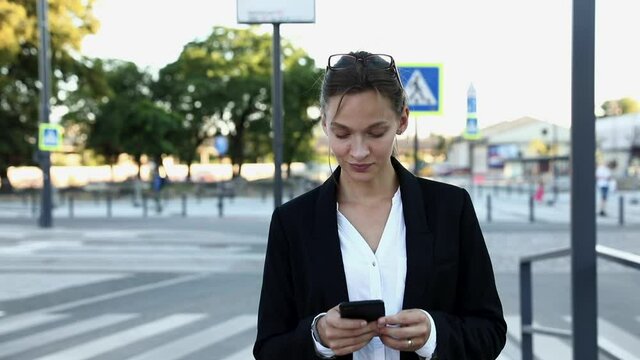 Successful business lady in formal clothing walking on street and browsing internet on smartphone. Attractive woman with brown hair working on distance with help of modern device.