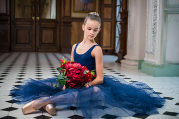 Portrait of a young ballerina on pointe shoes in a dark blue leotard and pointe shoes. Sits with a bouquet in his hands on the floor in a luxurious interior.