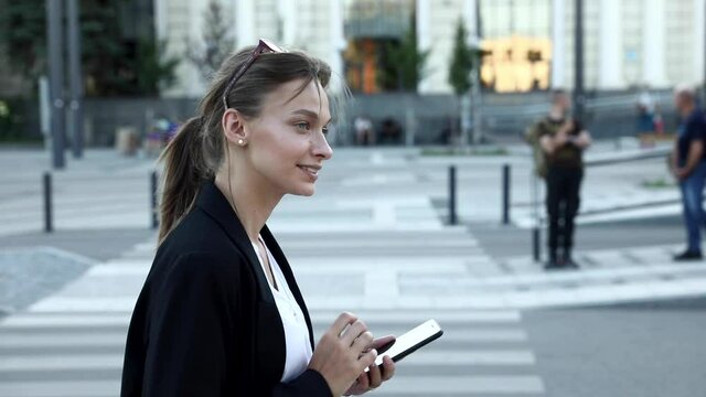 Side view of smiling brunette woman in casual clothing walking on street and using modern cell phone. Blur background of public transport. Concept of city life and technology.