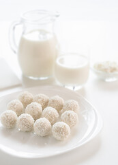 White sweet coconut truffles on a plate, milk glass and jug, cottage cheese in a bowl at defocused background, on a white table, in a high key.