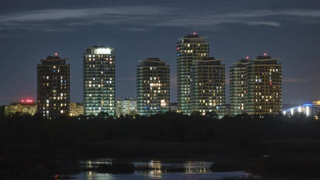 Time lapse from blue hour to night of Bucharest city skyline with tall buildings reflecting in Vacaresti Park Nature Reserve lake and apartment lights turning on and off.