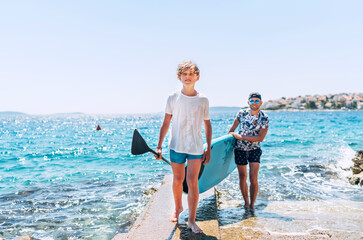 Blonde teenage son with father surfers smiling and carrying the stand-up paddleboard with a paddle on the bright sunny day noon. Active family summer vacation time near the sea concept image.