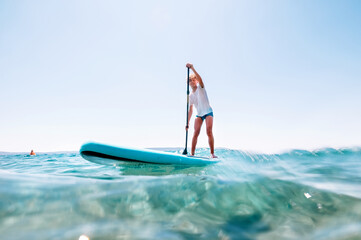 Under the water view angle to the smiling blonde teenager boy rowing stand up paddle board. Active...