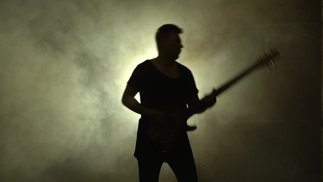 Silhouette of a young guy playing on the electric guitar on stage in a dark studio with smoke and neon lighting.