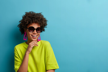 Fototapeta na wymiar Cheerful fashionable curly ethnic woman holds chin, wears trendy sunglasses and green t shirt, has happy summer mood, poses for magazine cover, isolated on blue background with copy space aside