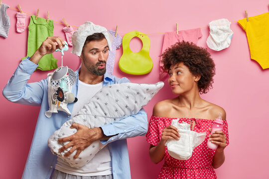 Funny Father Tries To Sooth Newborn, Shows Nipple And Talks To Baby, Wears Diaper On Head, Happy Cheerful Mother With Nappy And Feeding Bottle In Hands. Glad Young Parents Watch Their Baby Sleeping.