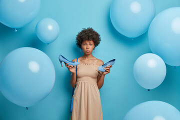 Upset woman got calluses while wearing high heel shoes, wears cocktail dress, has bad mood, tired after party, isolated on blue background decorated with inflated balloons. Women clothes collection