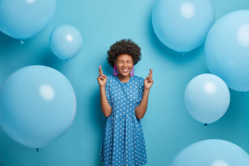 Happy celebration concept. Positive hopeful birthday girl crosses fingers, makes wish, believes all her dreams come true dressed in polka dot dress in one tone of background. Inflated balloons around