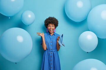 Fototapeta premium Crazy emotional curly woman looks happily at camera, happy to get heeled shoes as present from husband, dressed in everything blue, inflated balloons around. People, dressing and partying concept