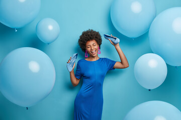 Fototapeta premium Pretty curly woman chills at party, dances happily, keeps hands raised with shoes, spends time in night club, takes off high heel shoes, poses against blue wall. Monochrome shot. Holiday, celebration