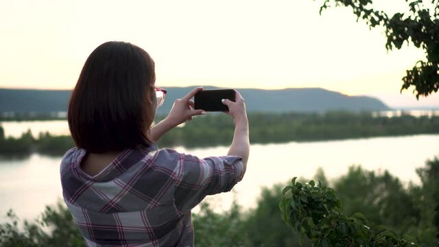 A young woman stands on a hill against the background of the river and mountains and photographs the landscape. The girl takes pictures in nature.