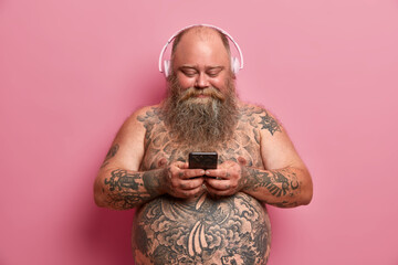 Pleased plump man with naked tattoed body, big belly, listens music in headphones, holds cellular,...