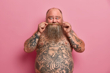 Funny bearded man touches mustache, stands naked with big belly, tattooed body, has fun and talks...