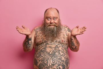 Photo of hesitant cheerful man spreads palms sideways, says I dont know, being happy and confused, has big tummy, tattooed body, doesnt know how to be fit and loose weight, isolated on pink wall - obrazy, fototapety, plakaty