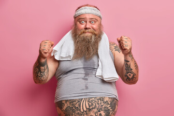 Funny overweight man being sweaty after intensive cardio, raises clenched fists, dressed in...