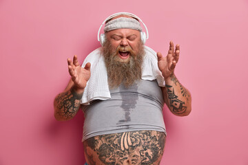 Emotive chubby hipster listens music in headphones, sings song loudly, dressed in sports wear, has...