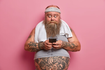 Shocked bearded man in sport outfit, takes break after physical exercises at home, holds smart...