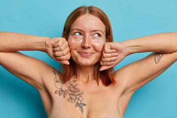 Beauty, cosmetology and rejuvenation concept. Smiling charming woman keeps hands on freckled face, wears transparent collagen patches, has anti aging eye therapy, freshed clean skin, tattoo on body