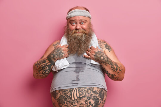Photo of funny blue eyed man has big belly, tattooed body, busy doing fitness exercises, burns calories after eating fast food, isolated on pink background. Plus size guy leads healthy lifestyle