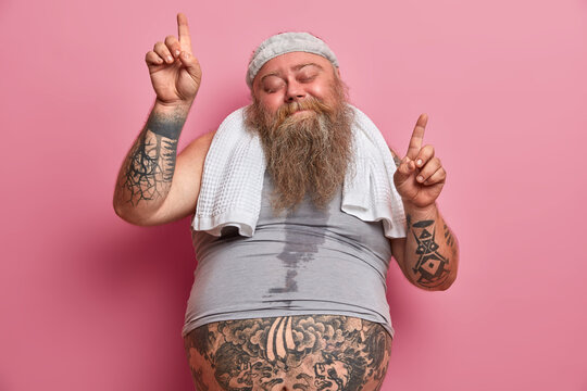 Obesity And Sport Concept. Joyful Overweight Man Dances Carefree Has Sweaty Body Tattooed Arms Points Upwards Isolated On Pink Background, Does Exercises At Home, Burns Calories After Eating Fast Food