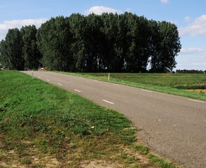 Partial view of an inland dike with a ramp on which a side road leads to the ridge of the dike. It protects the farmland behind it from the floods the river Meuse. There is a grove behind the dike © Gerfried