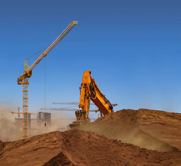 A dusty construction site with a yellow crane and an excavator bucket under a clear blue summer...