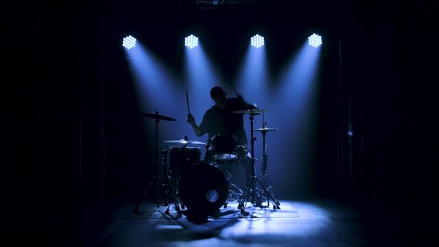 Silhouette drummer playing on drum kit on stage in a dark studio with smoke and neon lighting. Dynamic neon lighting effects. Performance vocal and musical band. Slow motion.