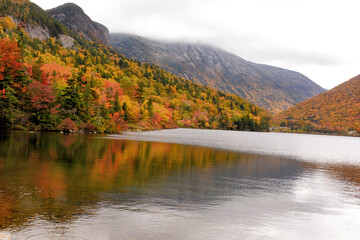 Autumn in White Mountains of New Hampshire. Sweeping view of Franconia Notch and colorful foliage from shore of Echo Lake. Dusting of snow on mountains and reflections of fall colors on lake.