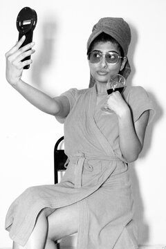 Attractive Young Indian Mexican Girl in Pink Bathrobe posing infront of Mobile Camera and Ring Light taking selfie smiling with eye glasses and cucumber on her eyes holding wine glass