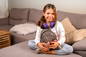 Little girl in musical headphones and a backpack is ready to go to school. A European schoolgirl demonstrates positive emotions. Education and school days concept. Copy space