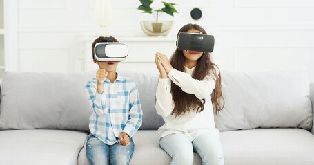 Caucasian small cute kids in VR glasses sitting on white sofa in living room. Little nice girl and boy, sister and brother playing at home, having virtual reality headset. Indoor. Augmented game world