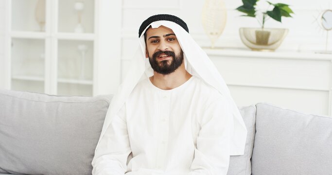 Portrait of handsome young Arabian man in kandura sitting on couch in living room and smiling to camera. Male Arab muslim in dishdasha on sofa at home. White interior Arabic millioner from Middle East