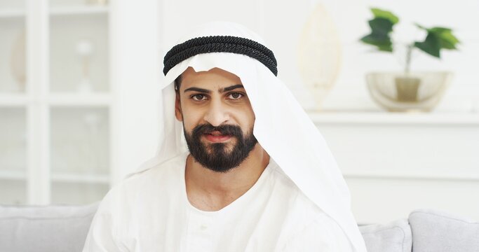 Close up of handsome young Arab man in kandura sitting on couch in living room and looking to camera with smile. Male muslim in dishdasha on sofa at home. Portrait of Arabic millioner from UAE.