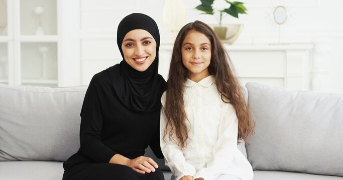 Portrait Of Beautiful Young Arabic Woman In Black Hijab Looking At Her Pretty Cute Teen Daughter With Love. Cheerful Charming Muslim Mother In Headscarf And Girl Smiling To Camera. Sitting On Couch.