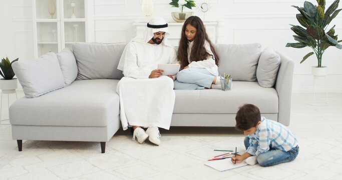 Arabian father in white kandura playing with small kids, daughter and son in living room. Muslim dad drawing and coloring pictures. Boy and girl with daddy painting at home and spending time together.